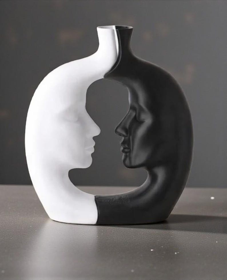 K&ouml;nnte beinhalten: Eine dekorative Vase in einem einzigartigen Design, das zwei sich gegen&uuml;berliegende menschliche Profile zeigt. Eine Seite ist wei&szlig;, die andere schwarz. Die Vase hat eine glatte Oberfl&auml;che und eine moderne &Auml;sthetik, geeignet f&uuml;r die Inneneinrichtung.