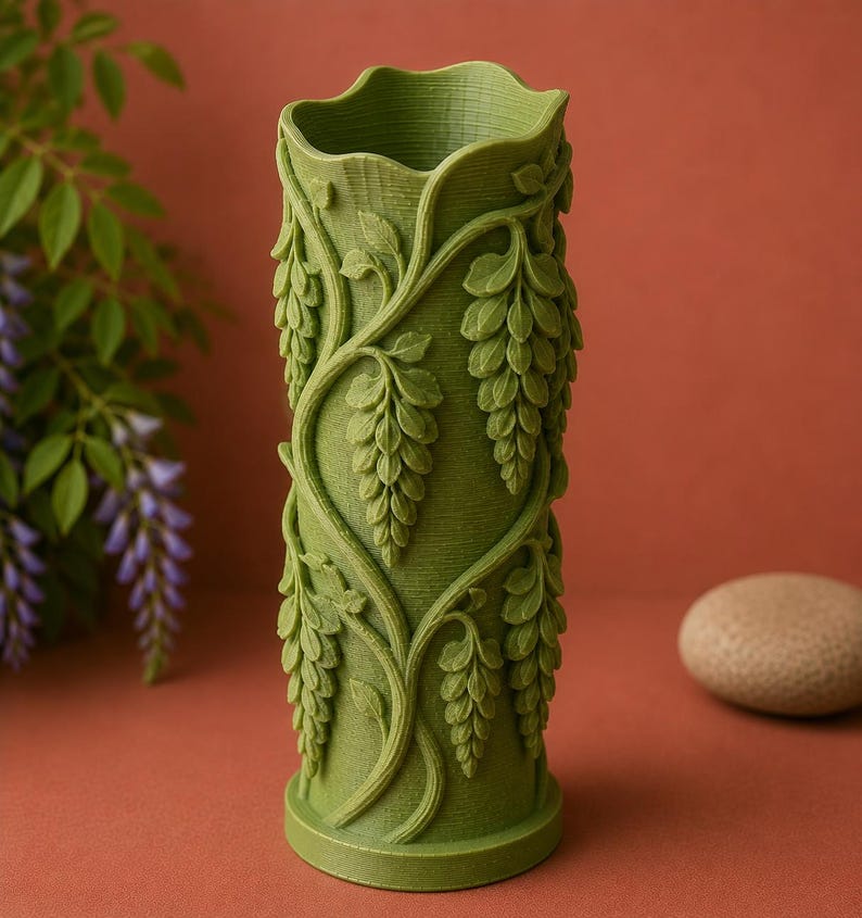 Puede incluir: Un jarr&oacute;n cil&iacute;ndrico verde con una superficie texturizada y un borde superior ondulado. El jarr&oacute;n est&aacute; adornado con un relieve detallado de vides de glicina y racimos de flores colgantes. El jarr&oacute;n mide unos 20 cm de alto.