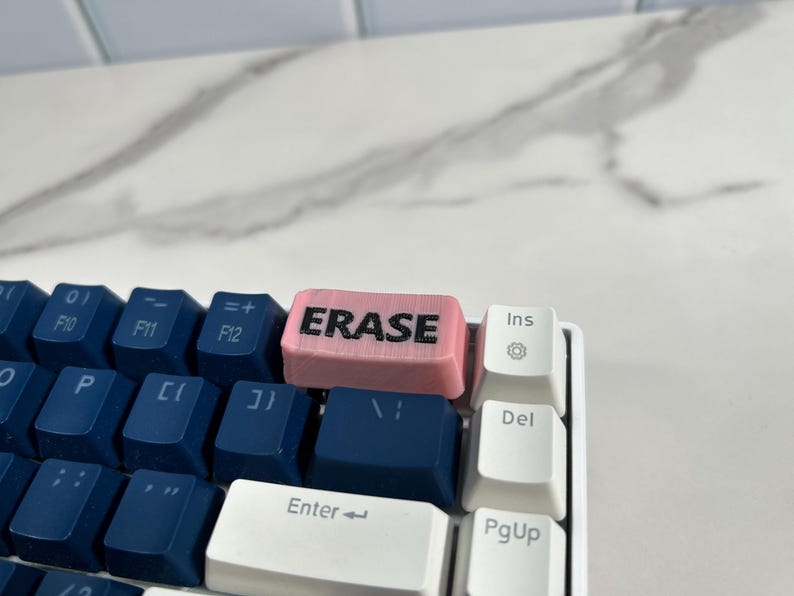 May include: A pink keyboard keycap with the word "ERASE" printed in black letters. The keycap is on a blue and white keyboard.