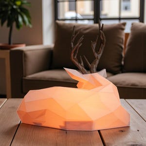 Puede incluir: Una l&aacute;mpara geom&eacute;trica con forma de ciervo con un c&aacute;lido resplandor anaranjado, apoyada sobre una mesa de madera. La l&aacute;mpara presenta un cuerpo blanco facetado y astas de color bronce. Es una l&aacute;mpara decorativa.