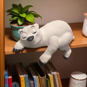 May include: A white polar bear figurine with black paws and nose is resting on a wooden shelf. The bear is lying down with its eyes closed. A small potted plant and books are visible in the background.