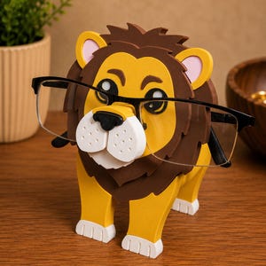 May include: A whimsical lion-shaped eyeglass holder. The holder is primarily yellow with a brown mane and accents, and white details. The lion is holding a pair of black-framed eyeglasses. The holder is designed to sit on a desk or table.