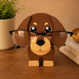 K&ouml;nnte beinhalten: Ein braun-beiger Dackel-Brillenhalter mit gro&szlig;en, cartoonartigen Augen und Ohren. Der Halter tr&auml;gt eine Brille mit schwarzem Rahmen. Der Halter steht auf einer Holzoberfl&auml;che. Eine Topfpflanze und ein Buch befinden sich im Hintergrund.