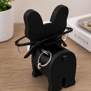 May include: A black and white bulldog-shaped desk organizer. The organizer holds eyeglasses and rings. The bulldog has a small drawer for storage. The glasses are black-framed. The rings are silver. The organizer is on a wooden surface.