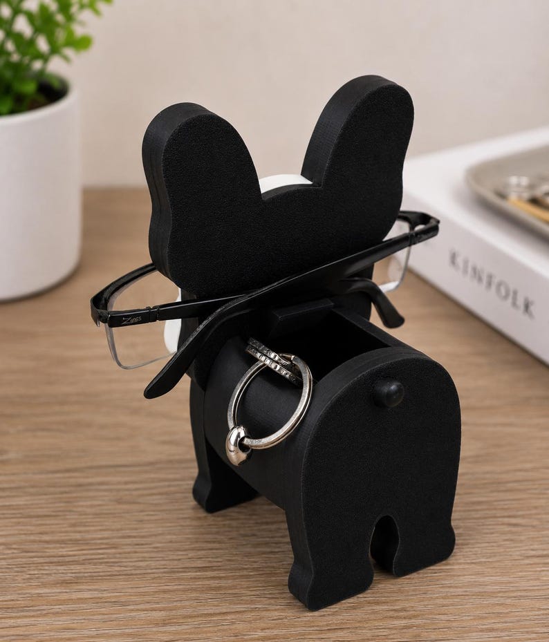 May include: A black and white bulldog-shaped desk organizer. The organizer holds eyeglasses and rings. The bulldog has a small drawer for storage. The glasses are black-framed. The rings are silver. The organizer is on a wooden surface.
