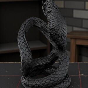 Puede incluir: Una escultura negra detallada de una serpiente en una pose enrollada. Las escamas de la serpiente est&aacute;n intrincadamente talladas y su lengua est&aacute; extendida. La escultura est&aacute; sobre un fondo oscuro, resaltando su forma y textura. La obra es una pieza decorativa.