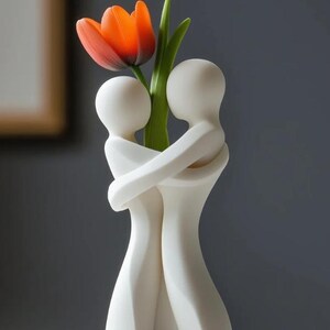 May include: A white ceramic vase in the shape of two embracing figures, holding a vibrant orange tulip with green leaves. The vase has a smooth, matte finish and is set against a gray background.