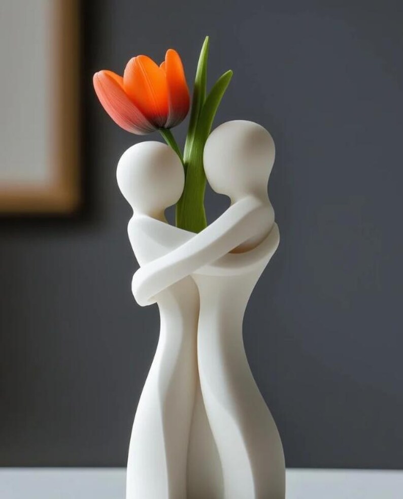 May include: A white ceramic vase in the shape of two embracing figures, holding a vibrant orange tulip with green leaves. The vase has a smooth, matte finish and is set against a gray background.