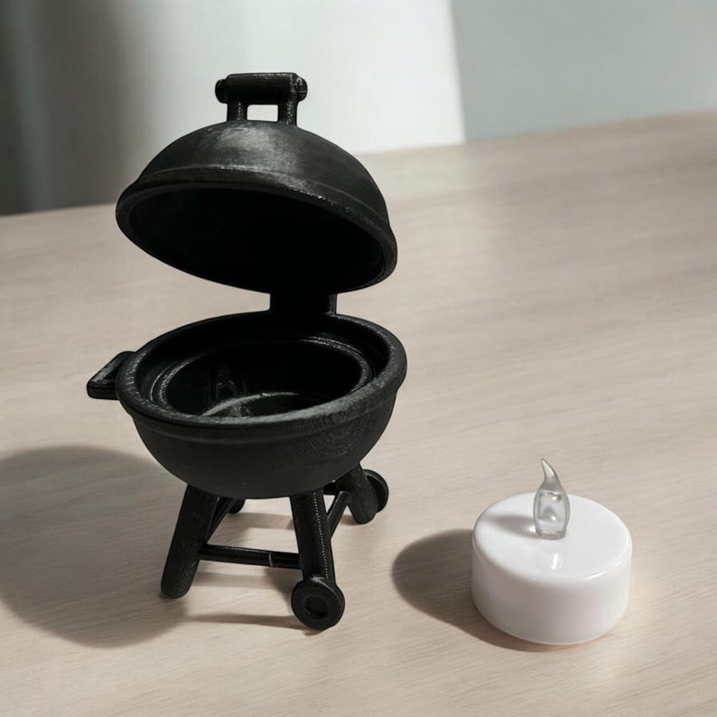 May include: A miniature black barbecue grill with the lid open, next to a small white LED candle. The grill has a round body, a handle on the lid, and sits on a wheeled stand. The candle has a clear, flame-shaped top.