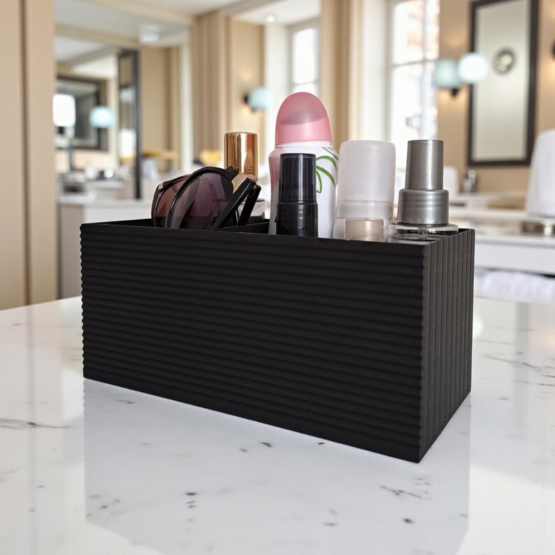 May include: A black, rectangular organizer with a ribbed texture, holding various beauty products. Items include sunglasses, lipstick, a spray bottle, and other cosmetic containers. The organizer sits on a white marble surface.