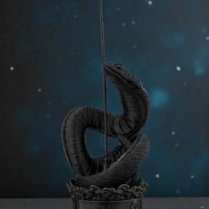 May include: A black incense holder in the shape of a coiled snake, with an incense stick emitting blue smoke. The snake is detailed with scales and sits atop a decorative base. The background is a dark blue with white dots.