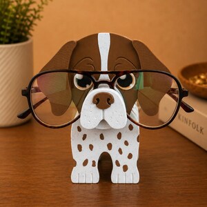 May include: A whimsical dog-shaped eyeglass holder. The holder is white with brown spots and a brown and white head. It is holding a pair of brown-rimmed eyeglasses. The holder is made of wood and is a decorative item.