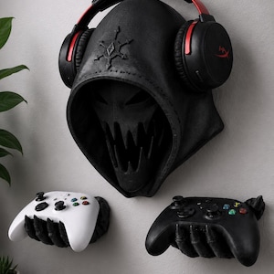 May include: A black wall-mounted display featuring a hooded skull mask with a star emblem, holding black and red over-ear headphones. Below, two black skeletal hand wall mounts hold a white wireless game controller and a black wireless game controller.