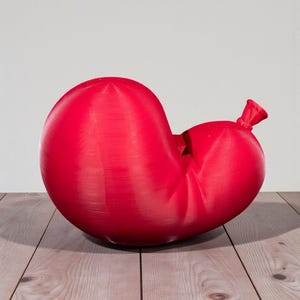 May include: A large, red, deflated balloon with a tied-off neck. The balloon is resting on a light-colored wooden surface, with a plain white background. The balloon has a textured surface.