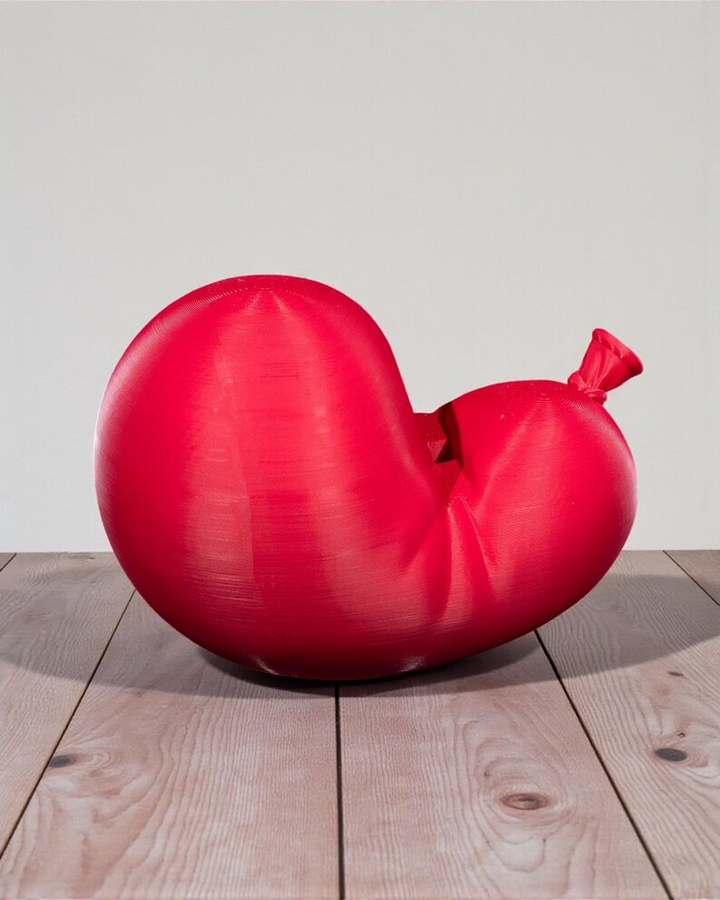May include: A large, red, deflated balloon with a tied-off neck. The balloon is resting on a light-colored wooden surface, with a plain white background. The balloon has a textured surface.