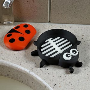 May include: A black and white ladybug-shaped soap dish with a white, slatted interior. An orange ladybug-shaped soap is next to the dish. The dish has two large, white eyes. The soap dish is on a speckled, light gray surface.