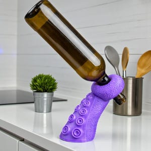 K&ouml;nnte beinhalten: Ein lila, oktopusf&ouml;rmiger Weinhalter, der eine braune Glasflasche tr&auml;gt. Der Halter hat Saugnapfdetails und einen Ring zur Sicherung der Flasche. Die K&uuml;chenumgebung umfasst eine kleine Pflanze und Utensilien.
