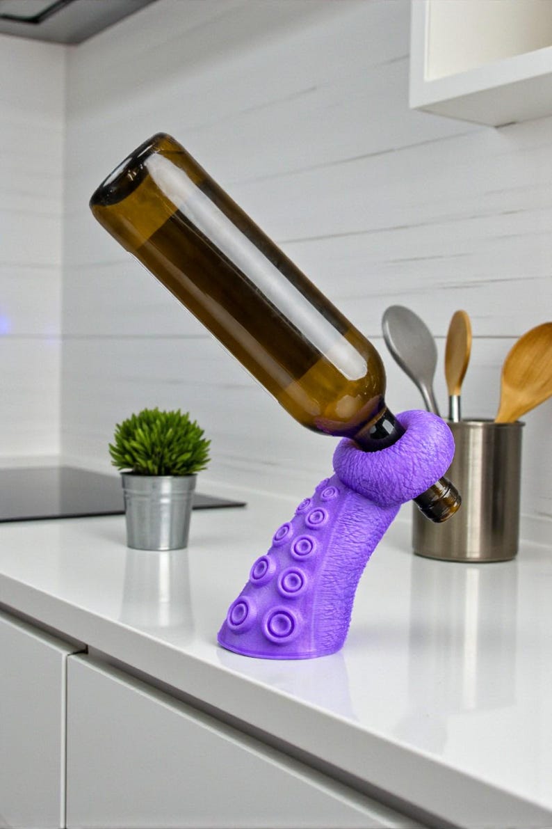K&ouml;nnte beinhalten: Ein lila, oktopusf&ouml;rmiger Weinhalter, der eine braune Glasflasche tr&auml;gt. Der Halter hat Saugnapfdetails und einen Ring zur Sicherung der Flasche. Die K&uuml;chenumgebung umfasst eine kleine Pflanze und Utensilien.
