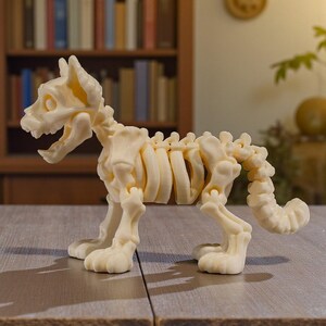 Puede incluir: Figura de esqueleto de perro de color crema con articulaciones articuladas. El esqueleto canino est&aacute; posicionado de pie, con la boca abierta. La figura se exhibe sobre una superficie de madera.