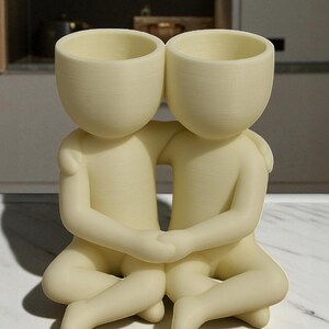 May include: A cream-coloured decorative planter featuring two figures embracing. Each figure has a rounded head with an open top, designed for planting. The figures are seated, with arms around each other, creating a unique and artistic home decor piece.