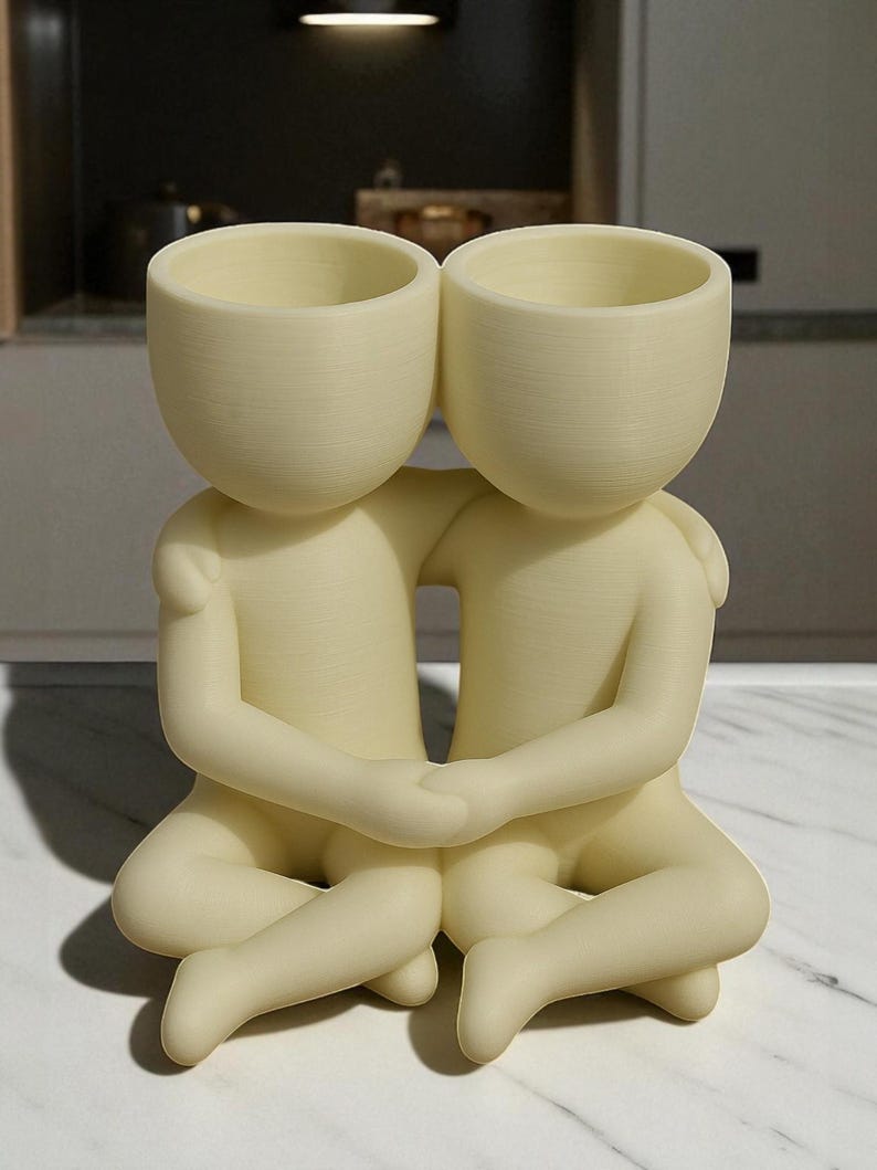 May include: A cream-coloured decorative planter featuring two figures embracing. Each figure has a rounded head with an open top, designed for planting. The figures are seated, with arms around each other, creating a unique and artistic home decor piece.