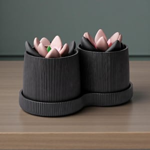 May include: Two black, ribbed ceramic planters with attached saucers. Each planter holds a decorative arrangement of pink, black, and green flower-shaped objects. The planters sit on a wooden surface.
