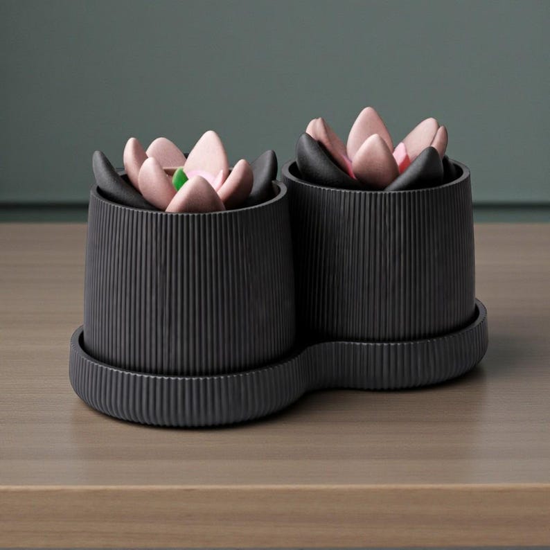 May include: Two black, ribbed ceramic planters with attached saucers. Each planter holds a decorative arrangement of pink, black, and green flower-shaped objects. The planters sit on a wooden surface.
