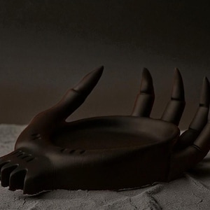 May include: A dark brown, hand-shaped decorative bowl. The bowl has a textured surface and is designed with open fingers and a shallow interior. The bowl is resting on a textured surface.