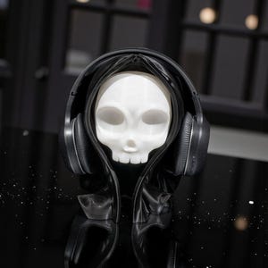May include: A white skull figure wearing a black hooded cloak and black headphones. The skull has hollow eye sockets and a small nose. The headphones are over the hood. The background is dark, with a reflective surface.