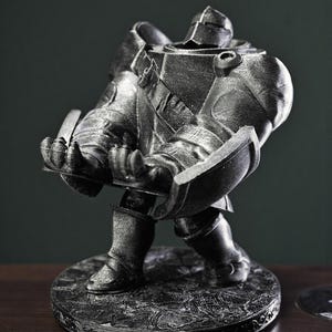 May include: A metallic silver figurine of a knight in full armor, holding a shield and a sword. The knight stands on a round, textured base with an ornate border. The statue is detailed and appears to be made of metal.