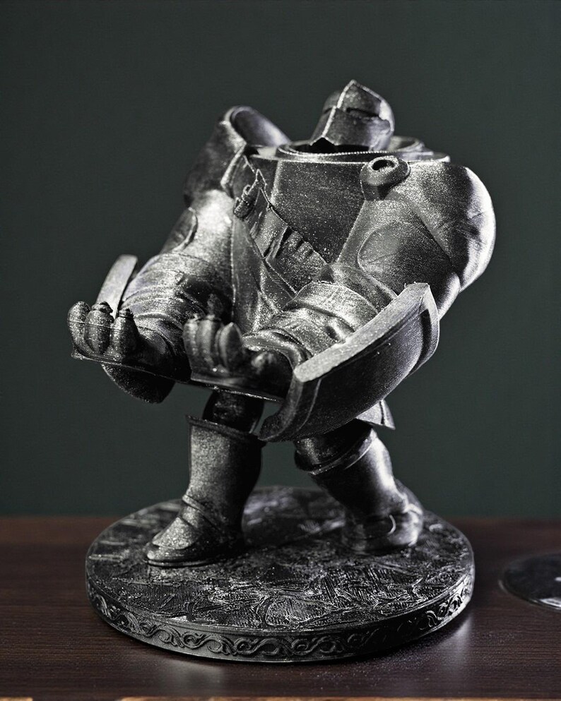 May include: A metallic silver figurine of a knight in full armor, holding a shield and a sword. The knight stands on a round, textured base with an ornate border. The statue is detailed and appears to be made of metal.