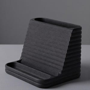 May include: A black desk organizer with a textured, ridged back and a small tray at the base. The organizer is made of a solid, matte material and is designed to hold pens, pencils, and other office supplies. The overall design is minimalist and modern.