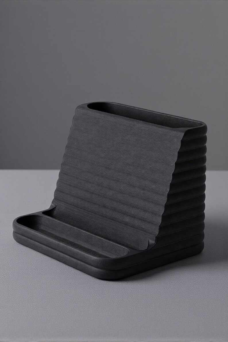 May include: A black desk organizer with a textured, ridged back and a small tray at the base. The organizer is made of a solid, matte material and is designed to hold pens, pencils, and other office supplies. The overall design is minimalist and modern.