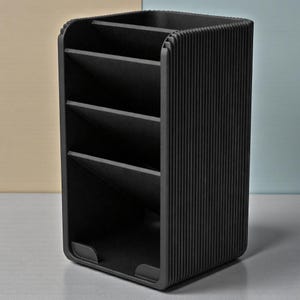 May include: A black desk organizer with multiple compartments. The organizer has a rectangular shape with rounded corners and vertical ribbed detailing on the sides. The interior features angled dividers to separate items. The organizer is designed for office or home use.