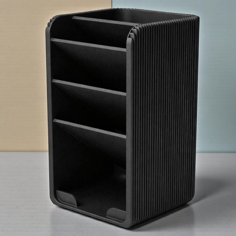 May include: A black desk organizer with multiple compartments. The organizer has a rectangular shape with rounded corners and vertical ribbed detailing on the sides. The interior features angled dividers to separate items. The organizer is designed for office or home use.