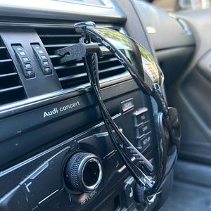 May include: A black car air vent clip with a pair of black sunglasses attached to it. The sunglasses are hanging from the clip and are resting on the car's dashboard. The car's dashboard has a black finish and features the text "Audi concert".