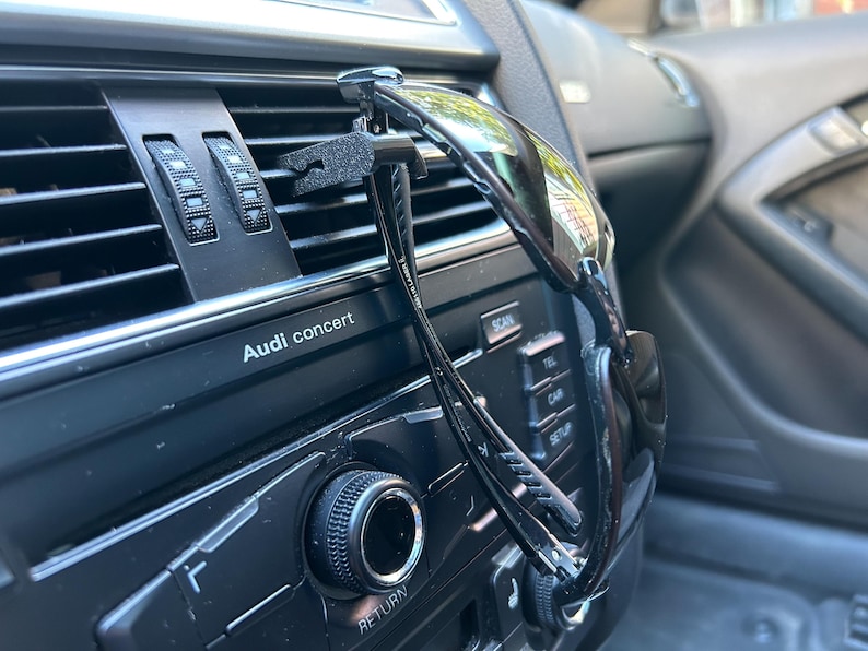 May include: A black car air vent clip with a pair of black sunglasses attached to it. The sunglasses are hanging from the clip and are resting on the car's dashboard. The car's dashboard has a black finish and features the text "Audi concert".