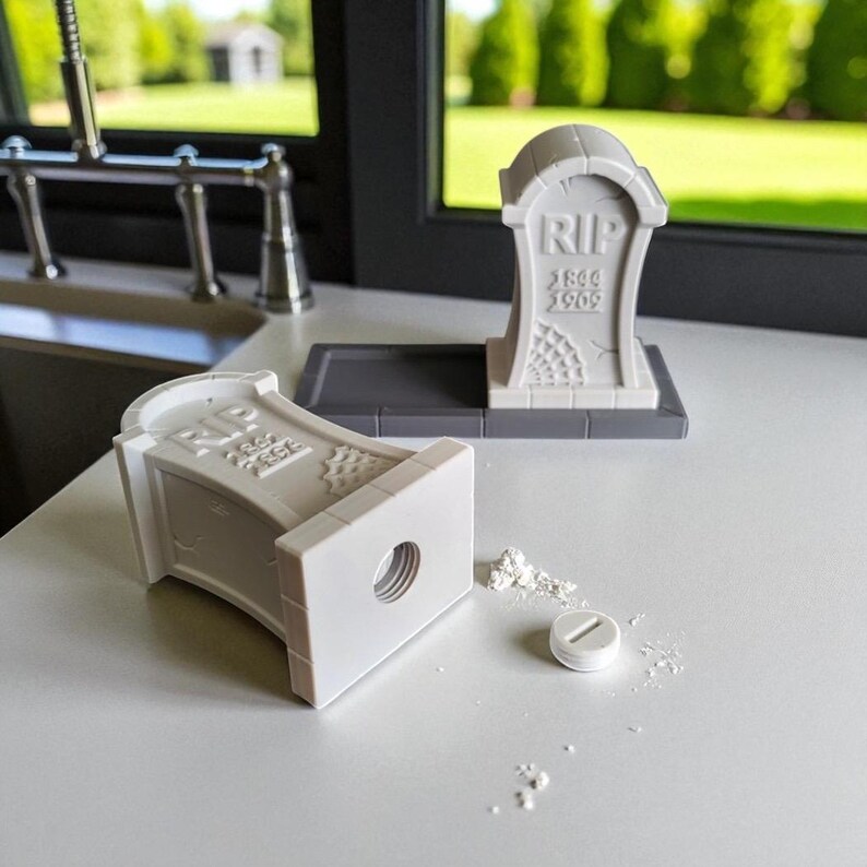May include: A gray tombstone-shaped pencil sharpener with the letters "RIP" and the years "1844 1909" etched on the front. A small white pill and pencil shavings are next to the pencil sharpener. The pencil sharpener is on a white surface.