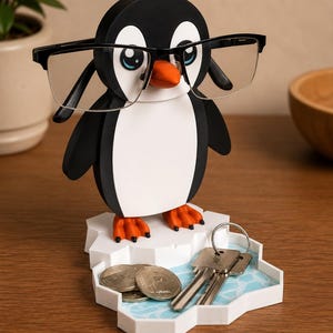 May include: A black and white penguin-shaped eyeglasses holder with a white base designed to look like an iceberg. The penguin has orange feet and a beak, and is wearing black-framed glasses. The base also holds keys and coins.