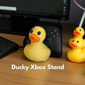 May include: A yellow rubber duck stand holding a black Xbox controller. The duck has an orange beak and black eyes. The text "Ducky Xbox Stand" is displayed below the duck. Additional rubber ducks are visible.