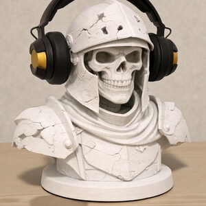May include: A white skull bust wearing a medieval helmet and armor, with black and gold headphones. The bust has a cracked, aged appearance and sits on a round base. The headphones have black earcups with gold accents.