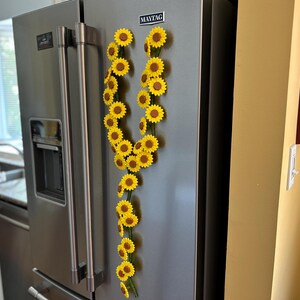 May include: A stainless steel refrigerator with a garland of yellow sunflowers attached to the door. The sunflowers have brown centers and green stems. The refrigerator is in a kitchen with white cabinets and a window.
