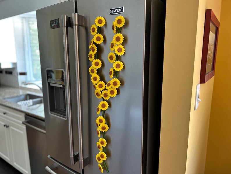 May include: A stainless steel refrigerator with a garland of yellow sunflowers attached to the door. The sunflowers have brown centers and green stems. The refrigerator is in a kitchen with white cabinets and a window.