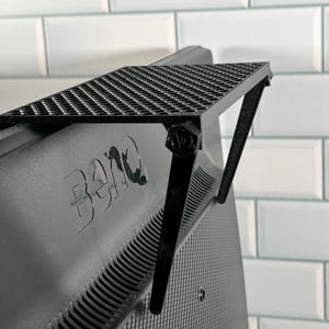 May include: Black monitor stand with a grid pattern on the top shelf. The stand is attached to the back of a black monitor with the BenQ logo visible.