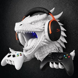 May include: A white dragon head wall mount featuring textured scales and an open mouth. The dragon wears black and orange over-ear headphones and uses its clawed hands to hold a white game controller on the left and a black game controller on the right.
