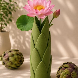 May include: A green vase shaped like a lotus flower holds a pink lotus blossom, a bud, and a green leaf. Two artichokes are on either side of the vase. The vase is on a light-colored surface.