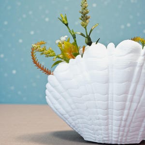 May include: A white seashell-shaped vase filled with colorful flowers and greenery. The vase has a textured surface and sits on a neutral-colored surface against a light blue background.