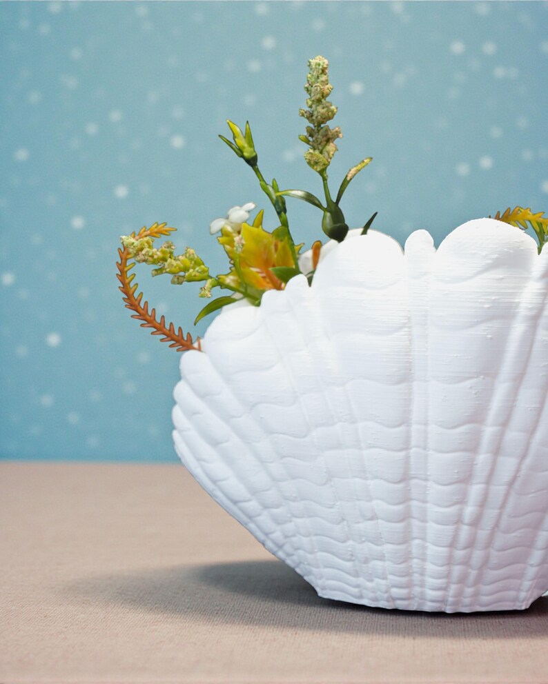 May include: A white seashell-shaped vase filled with colorful flowers and greenery. The vase has a textured surface and sits on a neutral-colored surface against a light blue background.