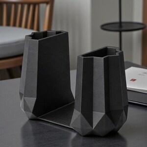 May include: A pair of black geometric desk organizers. The organizers have a faceted design with a matte finish. One section is a pen holder, and the other is a bookend. The organizers are made of a solid material and sit on a flat base.