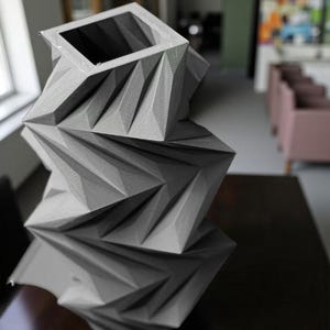 May include: A gray, geometric vase with a square opening and a faceted design. The vase is made of a textured material and sits on a dark surface. The design features sharp angles and a modern aesthetic.